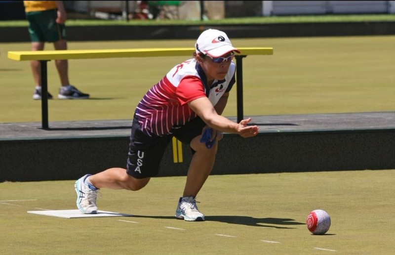 A lawn bowls player in a red, white, and navy “USA” uniform with a white cap and sunglasses crouches low on a mat as she releases a red bowl across a green, with a yellow bench and another player behind her.