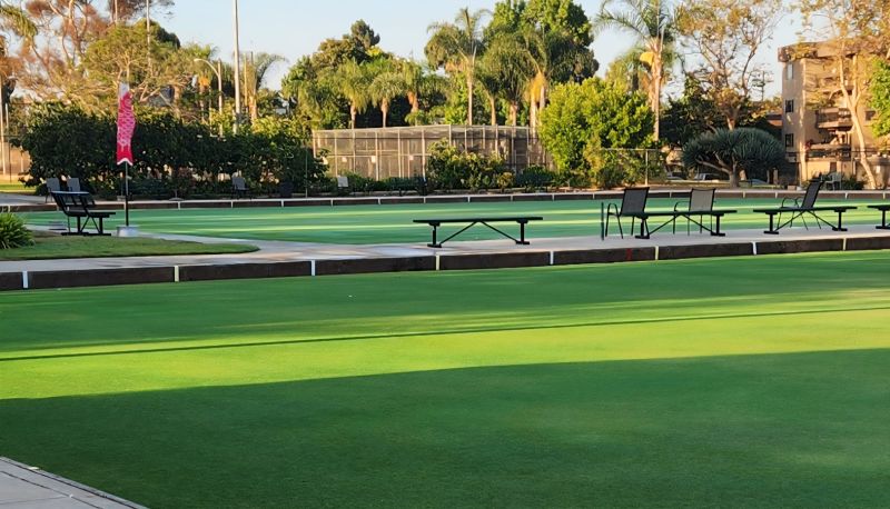 A manicured green lawn bowling green in warm sunlight, with benches and chairs along the edge, a pink flag, and palm trees and buildings in the background.
