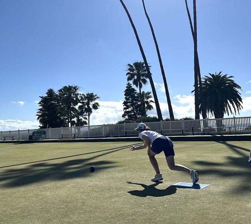 A lawn bowler in a white cap and navy shorts delivers a bowl across a sunlit green, with tall palm trees and a white fence behind her under a blue sky.