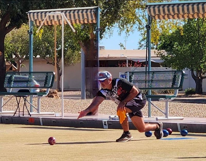 A lawn bowler in a black and white jersey, cap, and sunglasses crouches low as he delivers a bowl across a green, with shaded benches, other bowls, and desert landscaping behind him.